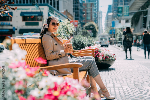 Happy pleased young asian woman chatting on mobile phone while listening music on headphones. smiling pretty female looks aside relaxing on bench with flowers in busy city street on sunshine day.