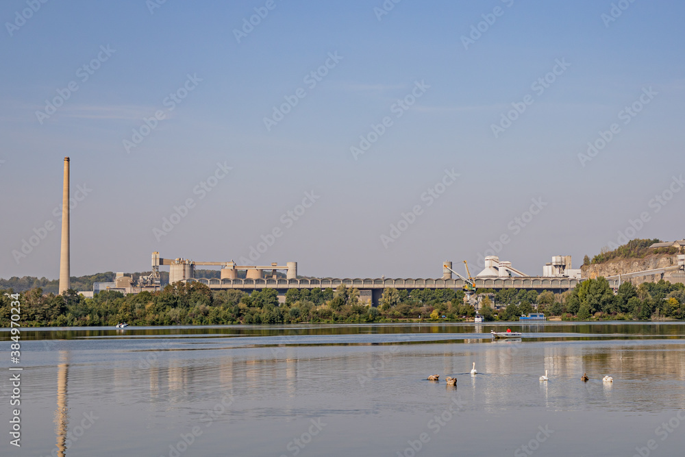 Maas River (Meuse) with slight reflection on its calm waters, swimming ducks and a small boat sailing with the old cement factory on the background, sunny summer day in South Limburg, the Netherlands