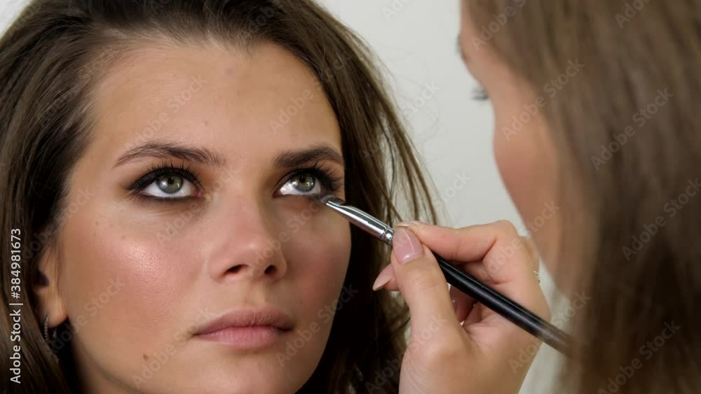 Attractive female face of a brunette well-groomed woman. Makeup artist applies eye shadows to the eyelid of the model eyes. Portrait of a beautiful young woman. Professional stylish makeup. Attractive female face of a brunette well-groomed woman. Makeup artist applies eye shadows to the eyelid of the model eyes. Portrait of a beautiful young woman. Professional stylish makeup.