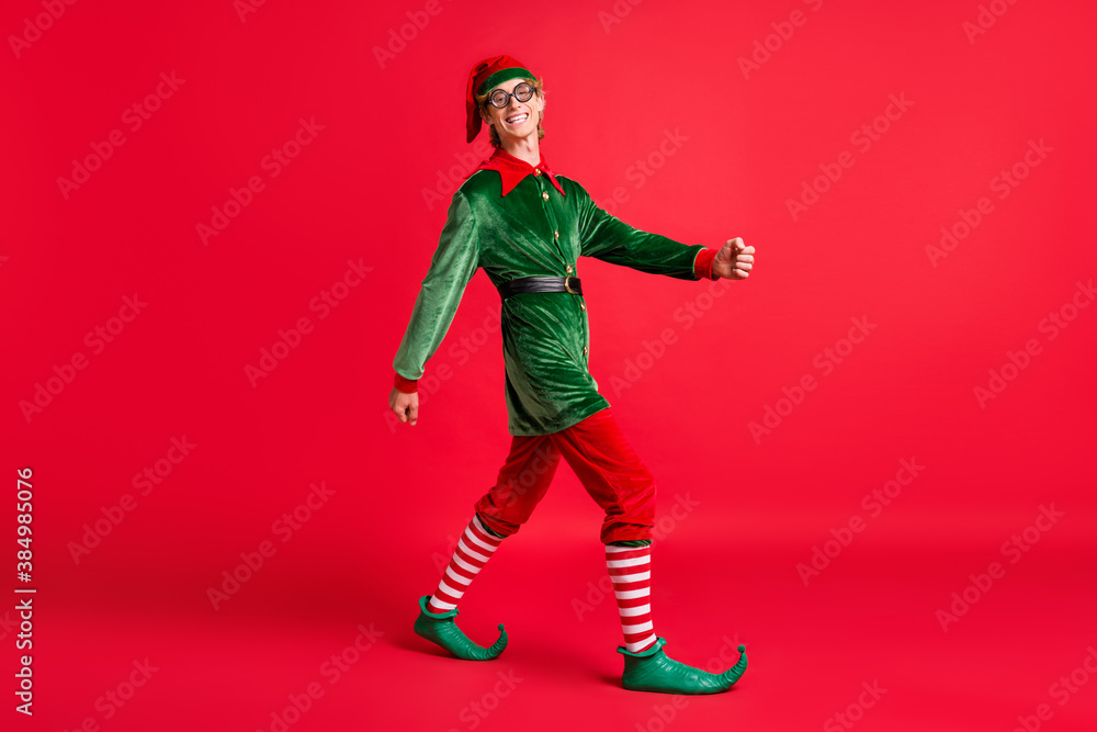 Full length profile side photo of positive elf guy go copyspace isolated over red bright color background Full length profile side photo of positive elf guy go copyspace isolated over red bright color background