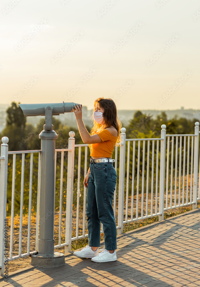 Young brunette woman looks through binoculars in public park while wearing a surgical mask