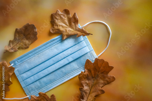 Fotografia A medical protective mask and yellow dry oak leaves lie on a bright autumn background
