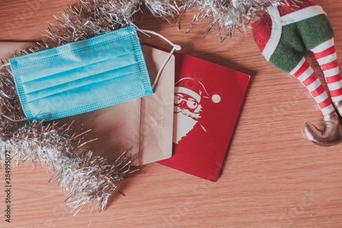 Una tarjeta roja de navidad con un Santa Claus blanco al lado de un espumillón plateado y una mascarilla quirúrgica azul photo
