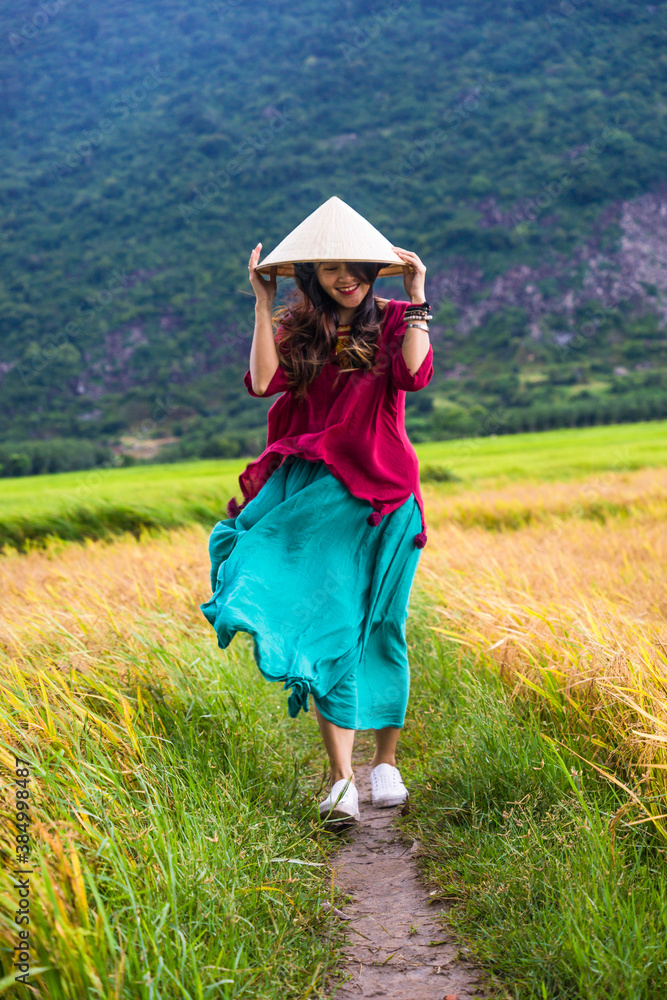 Vietnamese girl in dark red and bottle green traditional costume dress with conical hat walking on yellow and green rice field.