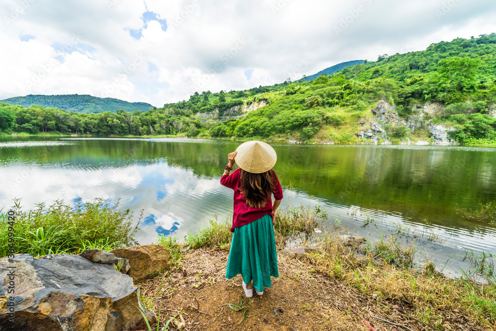 Vietnamese girl in dark red and bottle green traditional costume dress with conical hat standing in front of Ba Be lake