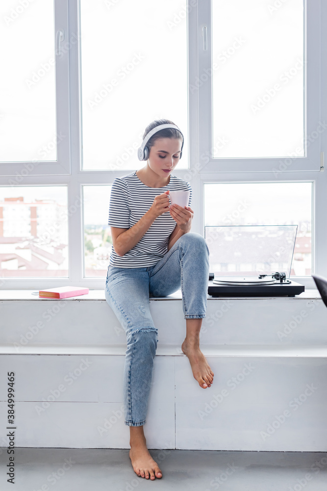 Barefoot woman in headphones holding cup while sitting near book and vinyl player on windowsill