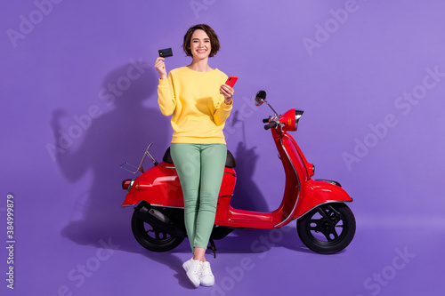 Full length body size view attractive cheerful glad girl sitting on bike using gadget bank plastic card order app shop isolated over bright vivid shine vibrant lilac violet purple color background