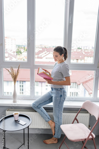 Young woman reading book near cup of tea and smartphone on coffee table