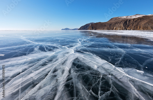 Beautiful winter landscape of frozen Baikal Lake with transparent smooth ice with cracks near the rocky shore in sunny cold day. Winter ice travel concept. Natural background