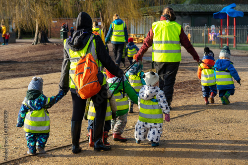 Nursery age children walking together with preschool teachers through a park wearing high visibility jackets