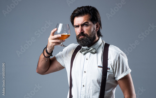 cheers. bearded man in suspenders drink scotch whiskey. brutal guy bartender wear bow tie. elegant male barman. handsome hipster drinking rum glass. party goer with alcohol. brandy or cognac