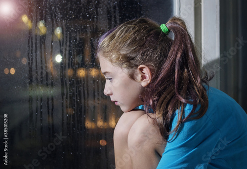 A brown eyed girl in blue t-shirt sitting on the window sill behind a foggy window with blurred night background