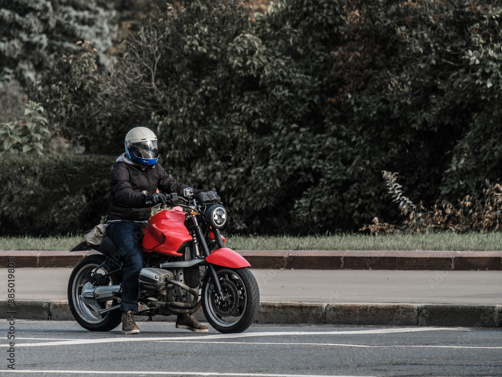 A motorcycle rider in a closed helmet sits on an unknown red sports bike during the day. The motorcyclist stopped on the asphalt with dense plants in the background. Freedom concept. A motorcycle rider in a closed helmet sits on an unknown red sports bike during the day. The motorcyclist stopped on the asphalt with dense plants in the background. Freedom concept.