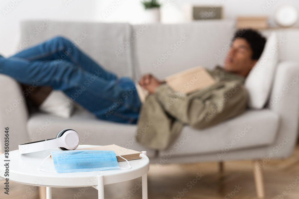 Blurred black teenage guy lying on sofa at home, selective focus of medical mask, book and headphones. Copy space Blurred black teenage guy lying on sofa at home, selective focus of medical mask, book and headphones. Copy space