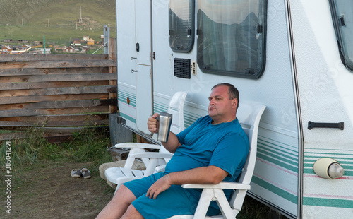 man is relaxing with a travel trailer RV. person on vacation with a mug in his hands. the concept of outdoor recreation. recreation water, long-distance travel and ecotourism. 