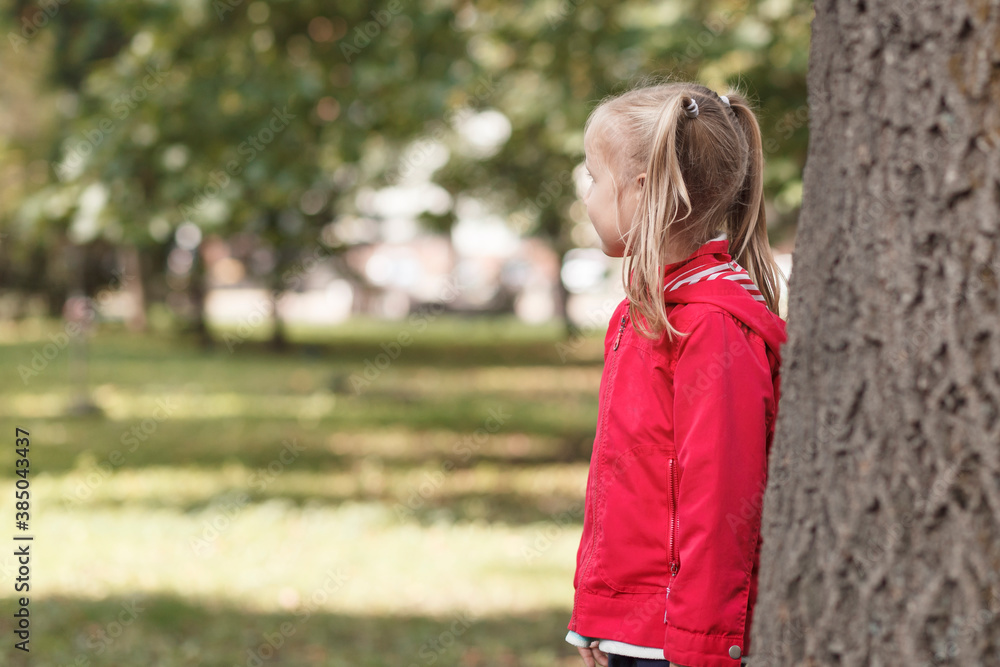 Little girl in a red jacket stands near a large tree in the park Little girl in a red jacket stands near a large tree in the park