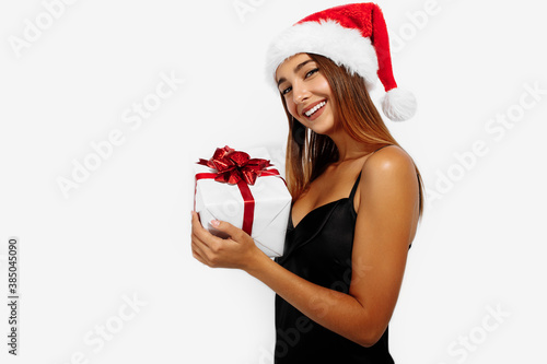Cheerful attractive brunette in a red Christmas hat and black dress, holding a gift box on a white background, Concept of Christmas, holiday