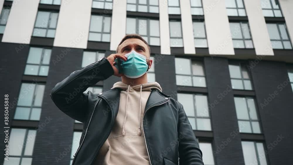Young stylish man wearing a protective mask talking by smartphone before modern building Young stylish man wearing a protective mask talking by smartphone before modern building