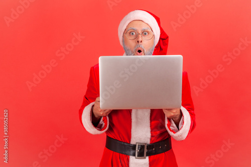 Photography Shocked surprised santa claus peeping behind laptop, looking at camera with big wondered eyes