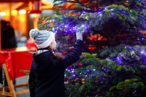Little cute kid girl having fun on traditional Christmas market during strong snowfall. Happy child enjoying traditional family market in Germany. Schoolgirl standing by illuminated xmas tree.