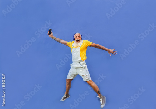 Cheerful hipster man jumping - Happy senior with smartphone and headphones enjoy music from playlist - Technology