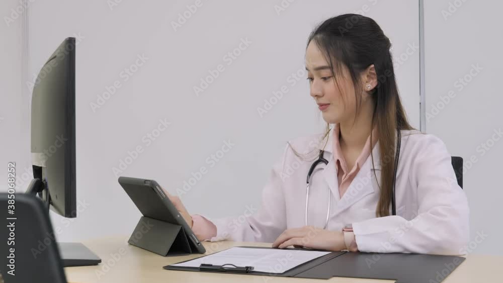 The young female doctor or nurse sits in the working room is using a tablet computer, and then she is looking at the camera with a beautiful smiling face. The young female doctor or nurse sits in the working room is using a tablet computer, and then she is looking at the camera with a beautiful smiling face.
