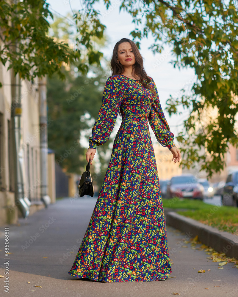 Elegant caucasian brunette woman with long wavy hair in long colorful dress with floral print walking at city street on autumn day