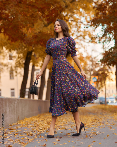 Elegant caucasian brunette woman with long wavy hair in midi purple dress with floral print walking at tree alley with colorful foliage photo