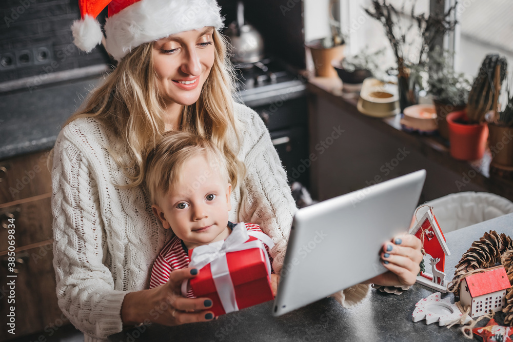 Mom in a Santa hat sits with a child at home, holds a tablet and makes a video conference call. Mother and son received a gift from their relatives and communicate by video call Mom in a Santa hat sits with a child at home, holds a tablet and makes a video conference call. Mother and son received a gift from their relatives and communicate by video call