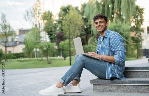 Portrait of happy Indian programmer using laptop computer, internet, working freelance project online, sitting in park. Asian student studying, learning language, using modern technologies outdoors