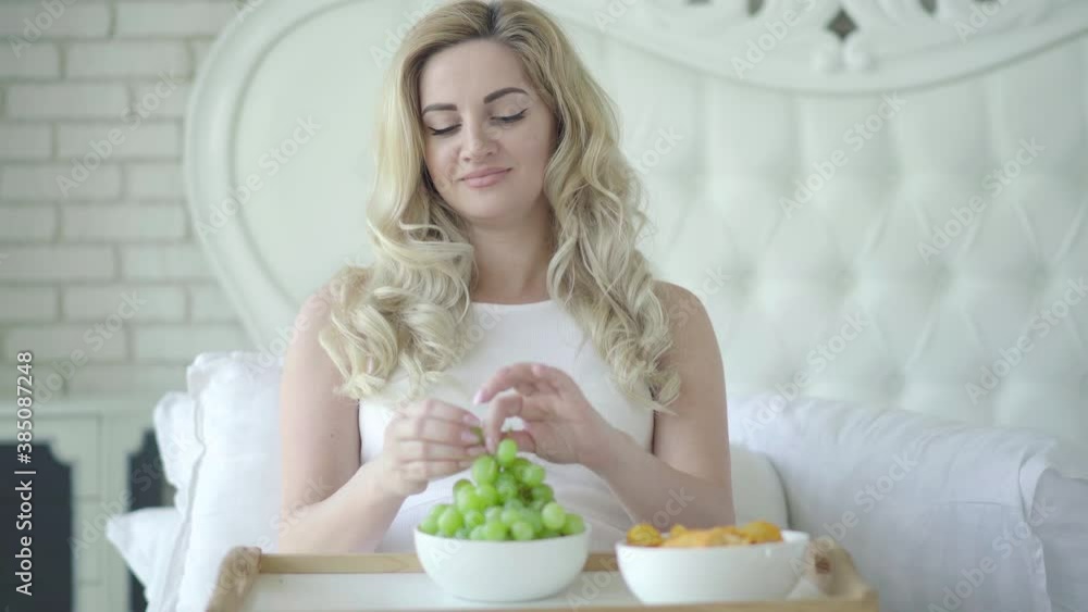 Portrait of positive beautiful pregnant woman eating healthful green grapes instead of harmful potato chips, and smiling caressing belly. Concept of healthy eating during pregnancy. Portrait of positive beautiful pregnant woman eating healthful green grapes instead of harmful potato chips, and smiling caressing belly. Concept of healthy eating during pregnancy.
