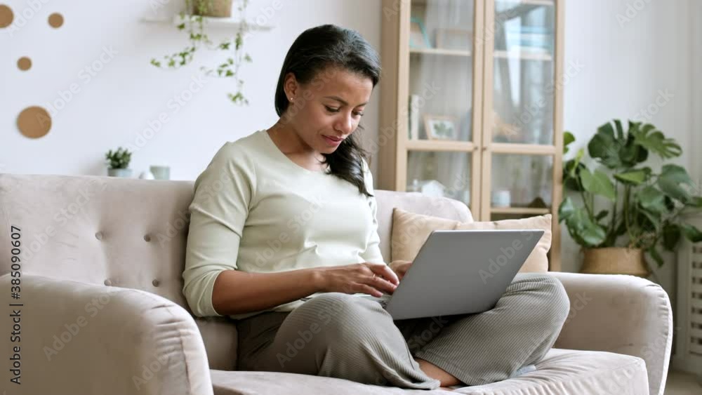 Pan shot of beautiful mixed-race woman sitting on sofa with laptop on her knees and typing message to friend Pan shot of beautiful mixed-race woman sitting on sofa with laptop on her knees and typing message to friend