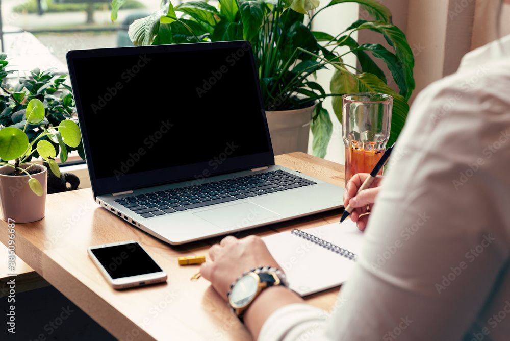 Young woman working on laptop and writing in notebook. Businesswoman at work in cafe near window with computer, phone and pen. Freelance and business. Office desk. Young woman working on laptop and writing in notebook. Businesswoman at work in cafe near window with computer, phone and pen. Freelance and business. Office desk.
