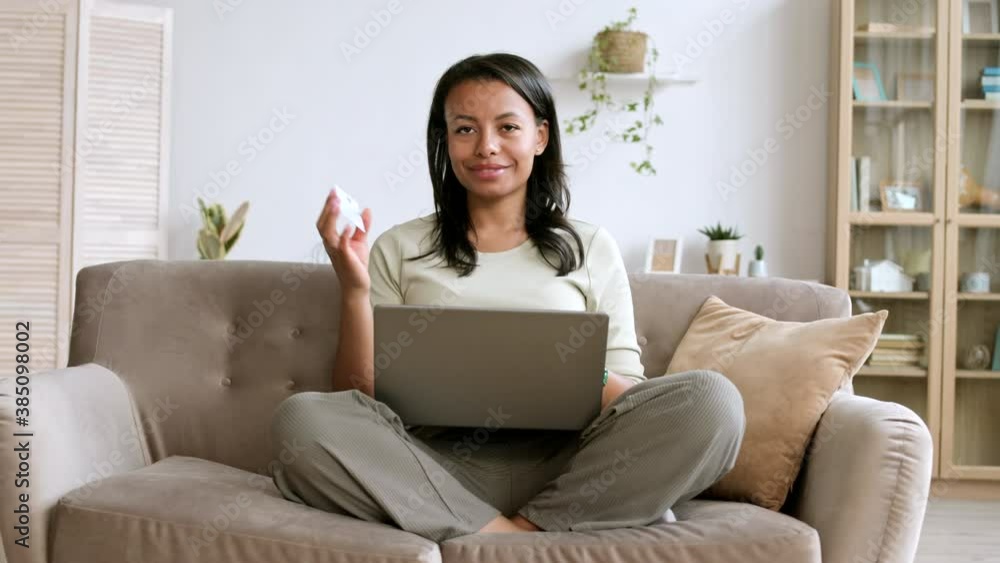 Medium shot of mixed-race woman with dark hair sitting on couch with laptop on her knees in living room, holding bank card in her hand and looking at camera Medium shot of mixed-race woman with dark hair sitting on couch with laptop on her knees in living room, holding bank card in her hand and looking at camera