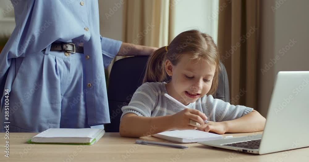 Girl sits at home, studies online remotely on e-learning, looks at laptop, does homework exercises, young woman mom older sister or teacher stand with child kisses rubs her nose on face of daughter Girl sits at home, studies online remotely on e-learning, looks at laptop, does homework exercises, young woman mom older sister or teacher stand with child kisses rubs her nose on face of daughter