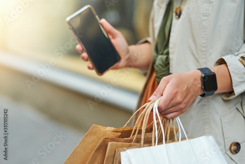 Cropped shot of a woman with shopping bags holding her smartphone while standing on city street outdoors