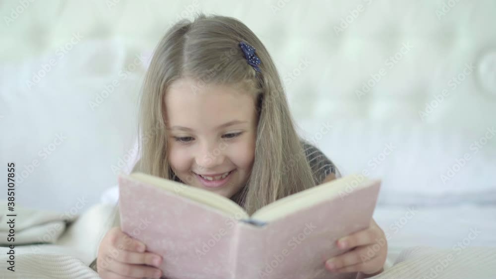 Portrait of cheerful Caucasian girl lying on bed and reading book. Pretty brunette child with brown eyes enjoying literature at home. Hobby and lifestyle concept. Portrait of cheerful Caucasian girl lying on bed and reading book. Pretty brunette child with brown eyes enjoying literature at home. Hobby and lifestyle concept.