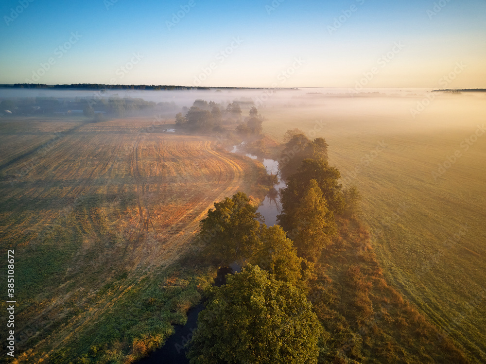 Small river with trees in meadow and field. Colorful misty morning dawn