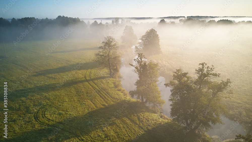 Autumn foggy sunrise aerial view. Small river with trees in meadow and field