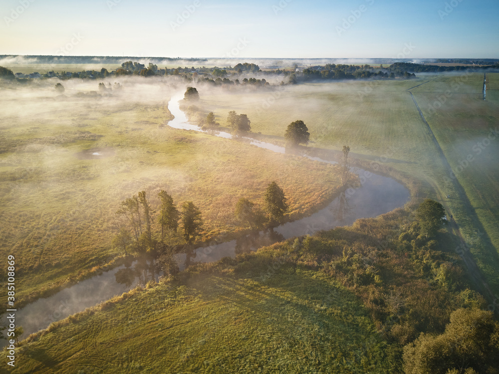Autumn foggy sunrise aerial view. Small river with trees in meadow and field
