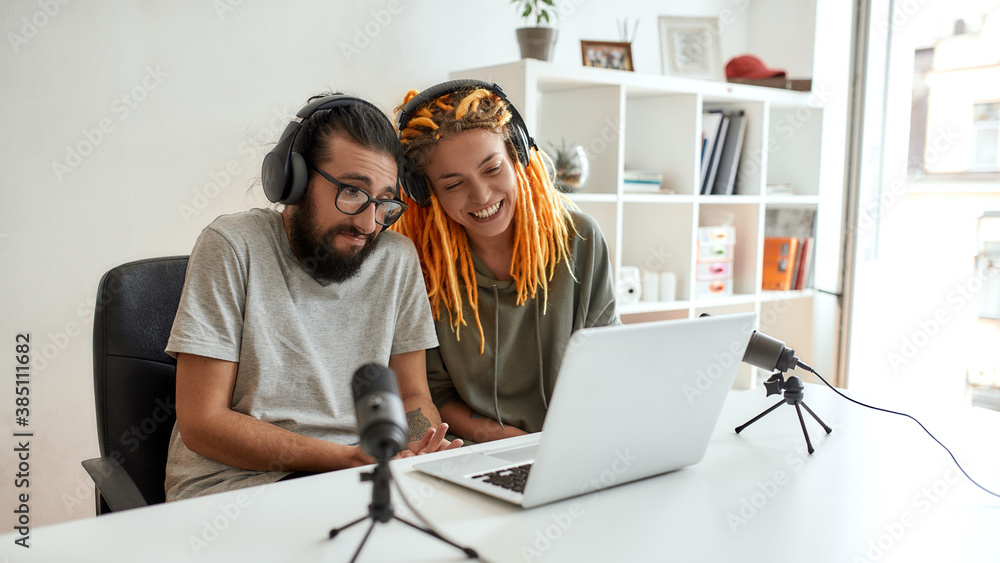 Couple of bloggers, young man and woman in headphones watching something on laptop, making faces while recording reaction video blog or vlog Couple of bloggers, young man and woman in headphones watching something on laptop, making faces while recording reaction video blog or vlog