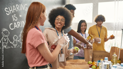 Happy young volunteers in gloves collecting, sorting food for needy people, Diverse team working together on donation project in charitable organization office