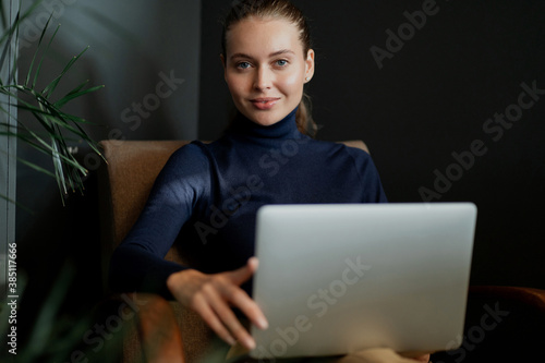 looking at the camera, a young confident woman is working on a laptop computer, embodying a new business task. stylish office clothes, hair pulled back, blonde Caucasian appearance