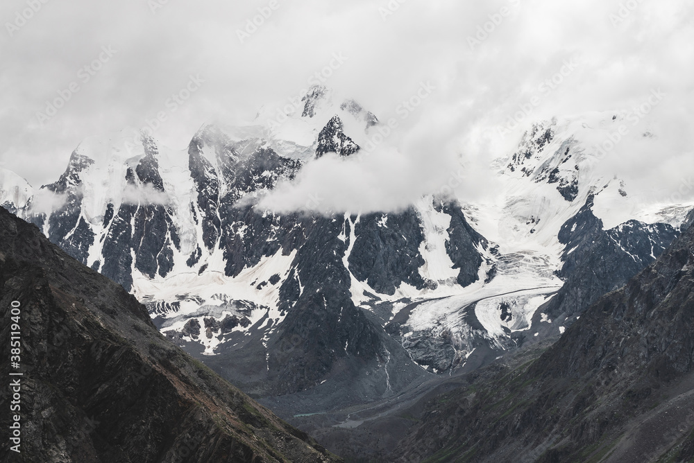 Atmospheric alpine landscape with massive hanging glacier on giant mountain. Big glacier tongue on mountainside. Low clouds among snowbound mountains. Cracks on ice. Majestic scenery on high altitude.