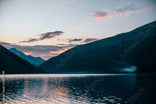 Vivid dawn sky is reflected in pure alpine lake near mountain silhouettes at sunrise. Colorful scenery with calm water of mountain lake. Beautiful tranquil landscape with water ripple in sunny morning © Daniil