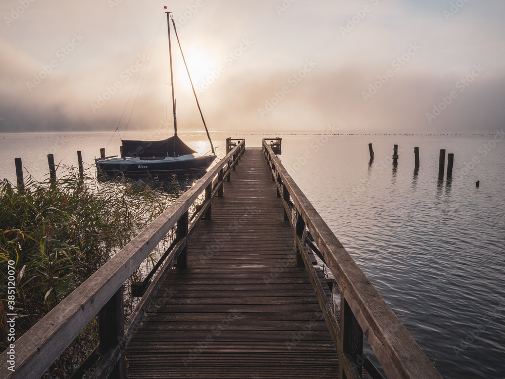  wooden jetty with a sailboat in the water of a lake in fog at sunrise