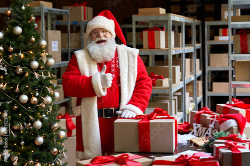 Happy old funny Santa Claus wearing costume holding Merry Christmas present gift box looking at camera showing thumbs up like standing in workshop warehouse. Fast xmas delivery concept. Portrait photo