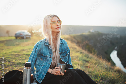 Portrait of young blonde girl on camping, wearing denim jacket and yellow glasses, holding steel cup with tea near thermos. Background of sunset.
