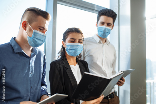 A group of collegues discuss business matters at the corridor of the office wearing medical masks to protect from coronavirus desease during global pandemic. Health safety at work concept.