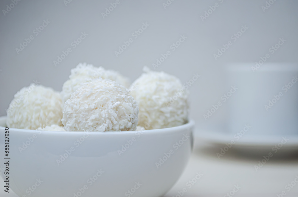 Round candies in a white plate covered with coconut shavings on a white background and with a Cup of espresso coffee. Round candies in a white plate covered with coconut shavings on a white background and with a Cup of espresso coffee.
